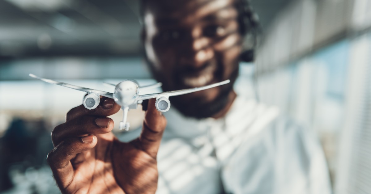 A smiling flight dispatcher holds a miniature airplane model between his fingers, making it appear as if it's flying.