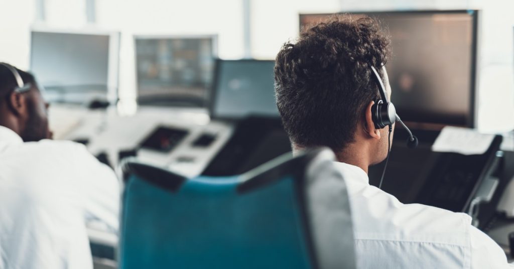 Two young flight dispatchers, wearing headsets, sit in the navigation room and monitor the screens before them.