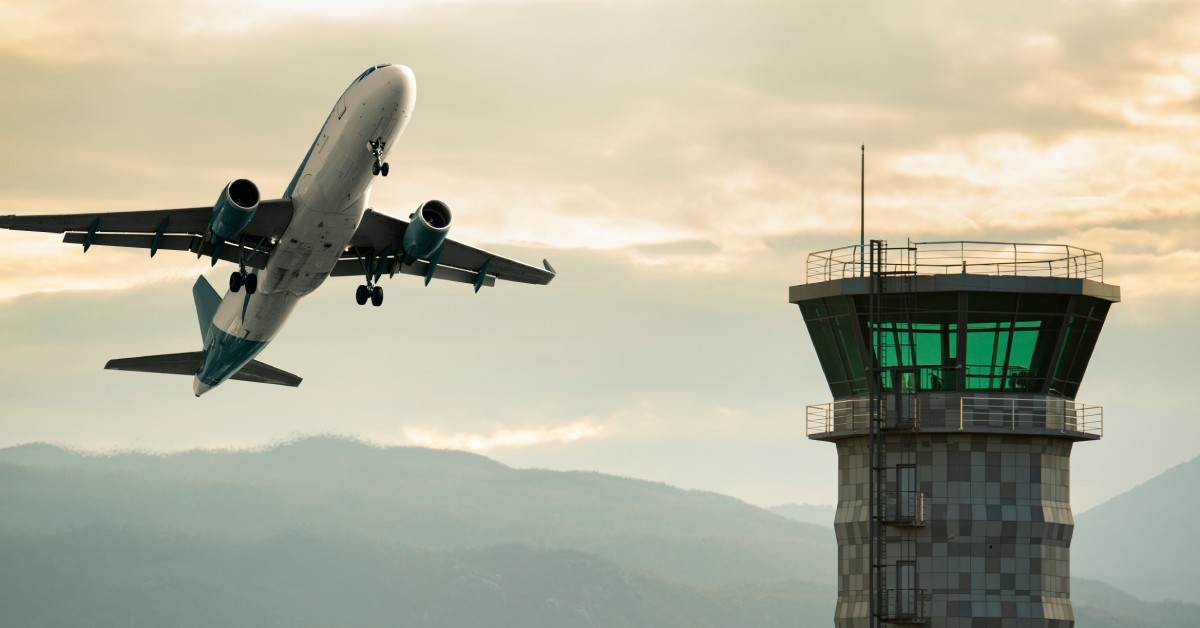 A white-and-blue passenger plane that's preparing to land soars past an air traffic control tower at sunset.