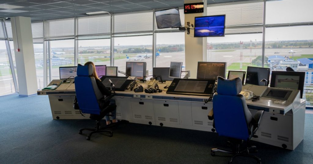 A woman sits in a desk chair in an air traffic control room. There is another desk chair next to her that is empty.