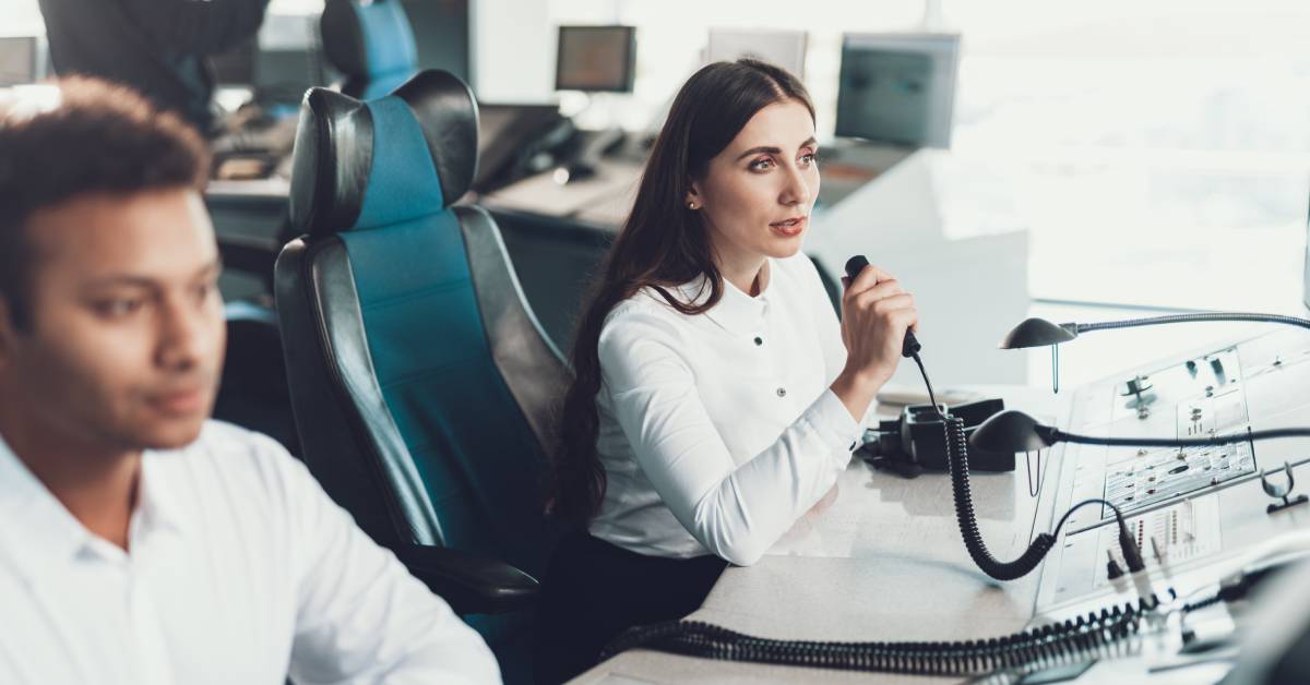 A woman wearing a white button-down shirt sits beside a man. She speaks into a microphone connected to a dispatch board.