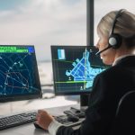 A woman in an air traffic control room wears a headset. She examines flight paths on a computer monitor.